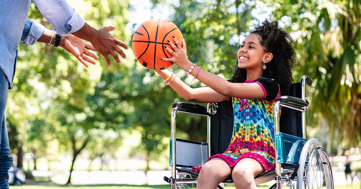 Menina negra em uma cadeira de rodas, sorrindo, com uma bola de basquete na mão